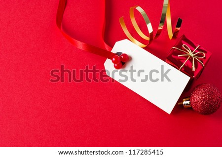 A blank Christmas gift tag, with red ribbon and artificial holly berries, on matte red background with shiny small gift box, glittery bauble, and gold foil spiral.  Copy space on tag and to it\'s left.