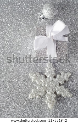 Overhead shot of three silver glittering Christmas ornaments on silver glitter background. Includes a bauble, a Christmas gift with white ribbon bow, and a star shaped snowflake.  Copy space to left.