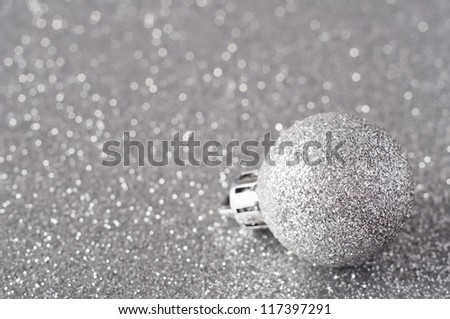 A single silver bauble, coated in glitter, resting on a silver glitter surface that softens into bokeh in the background.