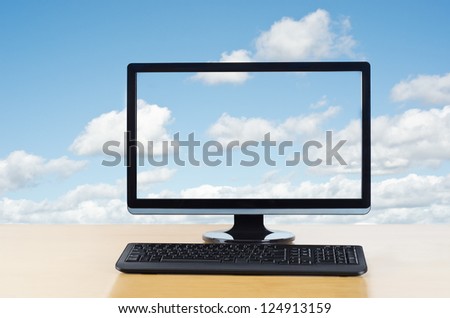 Conceptual image of cloud computing.  Computer monitor and keyboard on light wood table facing viewer,  in front of fluffy white clouds on a blue sky, which also flow unbroken through computer screen.