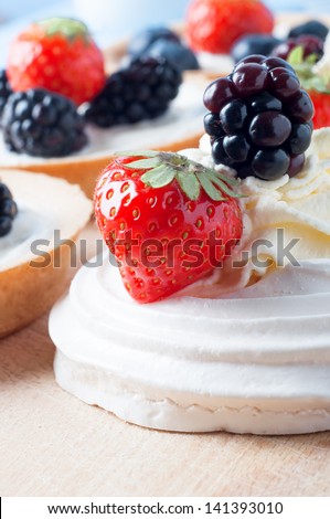 Close up of a meringue with swirls of fresh cream and berry fruits (strawberry and blackberry) on a wooden chopping board. In soft focus to it\'s side are fresh berry tartlets.