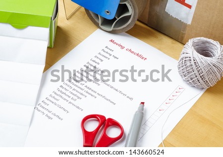 A house moving checklist on a table, surrounded by labels, packaging tape roller, scissors, red marker pen, a ball of string and a sealed box.  Some of the checkboxes have been ticked in red.