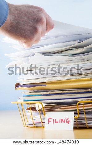 Business image of a male hand with blue shirt cuff visible, adding or removing document from tall pile in overflowing office filing tray.