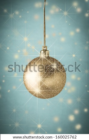 A gold bauble, layered with twinkling glitter and sparkling stars against vignetted turquoise blue background scattered with shining gold stars and and soft snowy orbs.