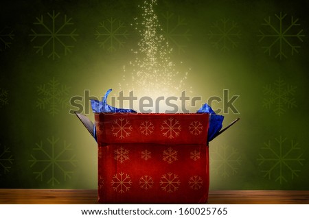 An opened red Christmas gift box with snowflake patterns on wooden table, emitting a magical warm bright glowing light and rising sparkling stars.  Green patterned fabric wallpaper effect background.