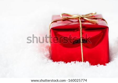 A single Christmas gift box in shiny red foil wrapping and tied to a bow with gold string ribbon, nestled in artificial white snow with copy space to the right.