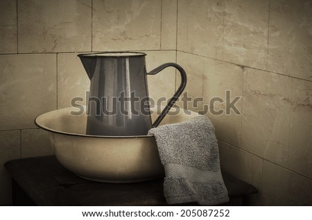 Blue enamel jug, wash basin and flannel on a wooden stand in a bathroom with tiled wall in background.  Vintage effect.