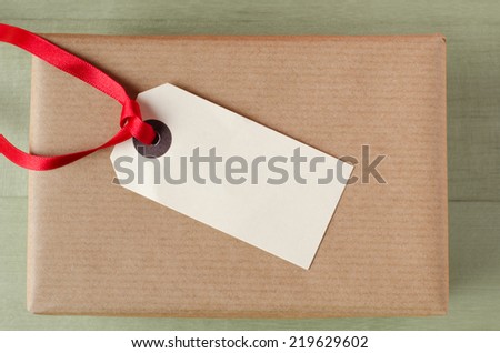 Overhead shot of a wrapped brown paper gift package on a green table. Topped with a parcel tag and red ribbon.  The blank label faces upwards to provide copy space for a message.
