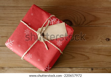 Overhead shot of a wrapped red Christmas parcel with snowflake pattern, tied with string.  Blank label faces upwards to provide copy space.  Set on an old, worn wooden table.