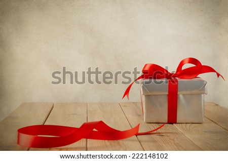 A Christmas gift box wrapped in silver paper and tied to a bow with a red satin ribbon, on a weathered old wooden table with copy space behind and above. Cut ribbon remnant to the side. Vintage style.