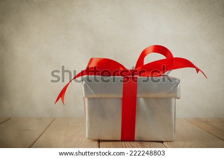 A Christmas gift box with closed lid, wrapped in silver paper and tied to a bow with a red satin ribbon.  Placed on a weathered old wooden table with copy space to left and above. Vintage style.