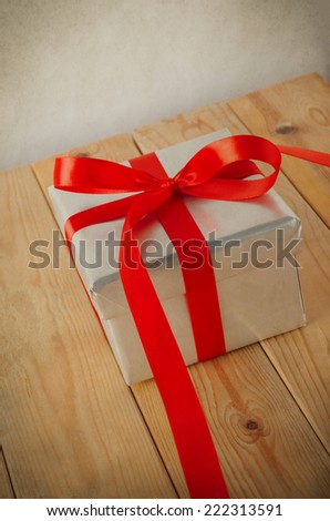 Tilted shot of a silver gift box, tied with red satin ribbon.  Ribbon end trails toward viewer.  Placed on wood plank table with parchment background.  Vintage style.