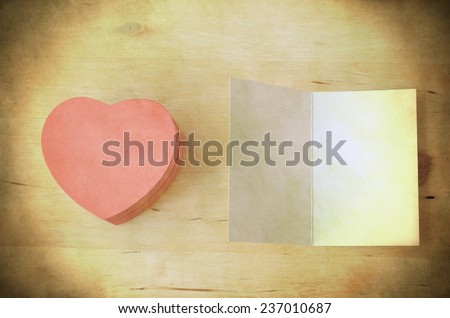 Overhead shot of a pale pink wooden heart-shaped gift box next to an opened blank card, providing copy space. Fresh retro style with light, bright yellowed toning and grunge style weathering.
