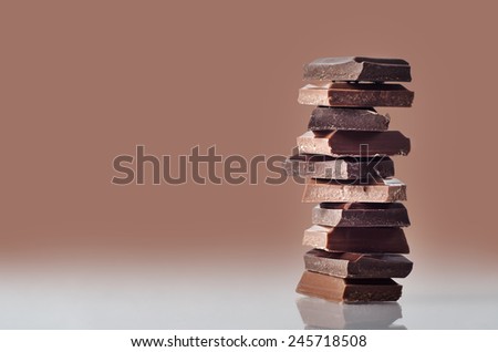 Eye level shot of chocolate pieces, broken from a bar and stacked in a column, standing on a reflective surface against a chocolate brown background which fades toward the surface.