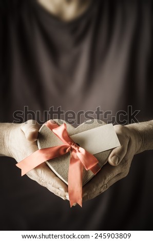 Male hands holding out a heart shaped gift box, tied to a bow with satin ribbon and blank facing front.  Man's body provides soft focus background.  Low saturation for retro feel.