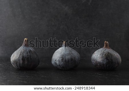A row of three whole figs on black slate against a black background with low lighting.