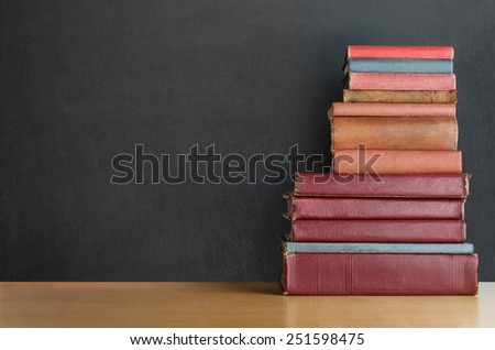 A pile of old, shabby, well used text books stacked in a pile on a wooden desk in front of a black chalkboard.  Copy space on left side.
