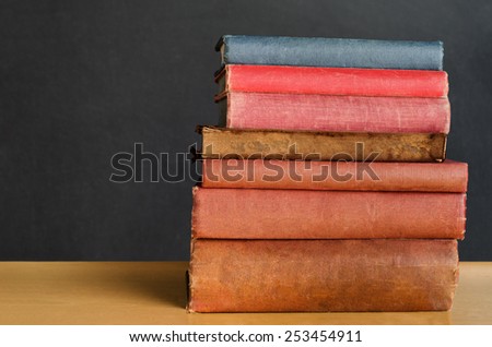 Close shot of a pile of old, shabby, well used text books stacked in a pile on a desk in front of a black chalkboard.  Copy space on left side.