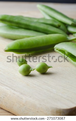 Close up of fresh, raw pea pods, scattered on a wooden chopping board, with three small peas extracted and placed in the foreground with stalks intact.