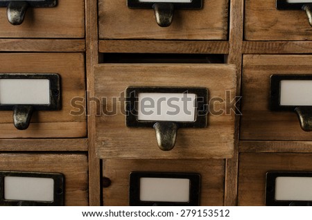 An old style wooden cabinet of library card index drawers with label holders and blank labels facing front.  One drawer in the middle is opened.