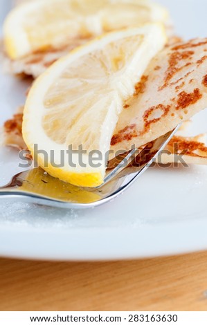 An English style pancake, topped with half a lemon slice and sprinkled with caster sugar, being lifted from white plate by shiny fork.