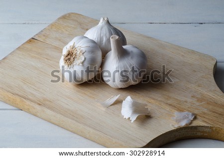 Angled elevated view of three garlic bulbs grouped on an old wooden chopping board with white painted wood plank kitchen table underneath.  Flakes of garlic paper scattered on board.