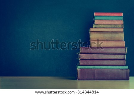 A pile of old, shabby, well used text books stacked in a pile on a light wood laminate desk in front of a black chalkboard.  Copy space on left side  Cross processed for retro or vintage effect.