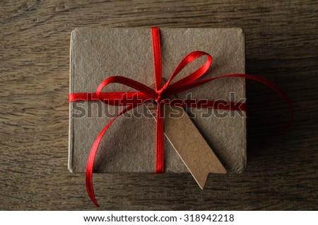 A plain brown cardboard gift box with blank vintage style notched gift tag, tied to a bow with thin red satin ribbon.  Overhead shot on oak wood table.