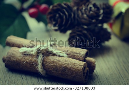 Cinnamon sticks, tied in a bundle with string on an oak table with Christmas holly, fir cones and a gold wrapped gift box in the background. Cross processed for retro or vintage effect.