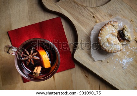 Overhead shot of a glass of mulled wine on red napkin, with partially eaten mince pie on wooden paddle board.  Light wood table below.
