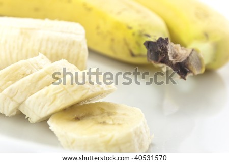 Close-up of banana slices on a white china plate, with remaining portion and two whole, unpeeled bananas in soft focus in the background.