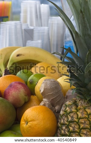 A selection of fruits laid out at a juice bar (stall), with cups and straws in soft focus in the background.  Includes bananas, oranges, apples, limes, lemons, ginger and pineapple.