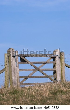 A wooden gate attached to a barbed wire fence, with bright blue sky beyond, on top of a grassy hill.
