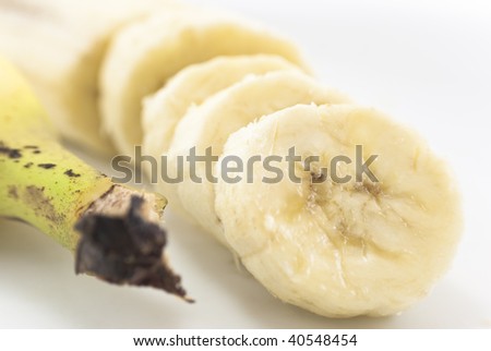 Close-up of sliced banana with remaining portion in soft focus behind.  To it\'s left is the top end (stalk) of an unpeeled banana.  White background.