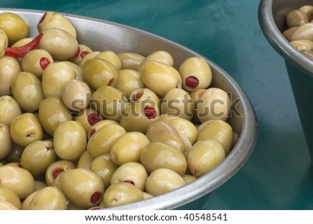 A metal bowl of green olives, stuffed with red chilli peppers, on a teal colored tablecloth on a market stall.