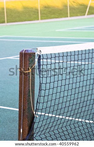 Tennis court net attached to rusting metal post with yellow and grey ropes.  Tennis court of green tarmac is visible.  Out of focus wire fence and shadowed grassy hill in background.  Winter daylight.