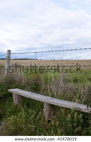A wooden seat in front of barbed wire at the edge of harvested farm land at the change of season from Summer to Autumn.  Poppies and power lines visible.  Cloudy blue sky.