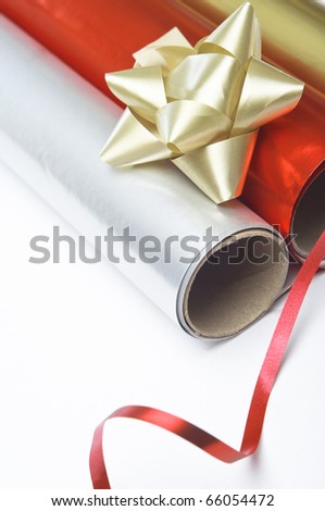 Close up, angled shot of gift wrapping paper rolls, rosette and ribbon on a white surface.