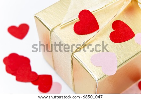 Close up (macro) of a gold gift box, tied and bowed with organza ribbon and scattered with pink and red paper heart confetti.  Background is off-white.