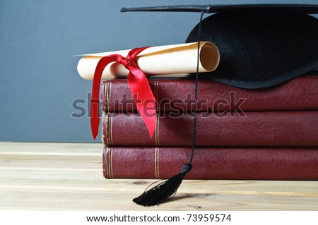 Graduation mortarboard and scroll tied with red ribbon on top of a stack of old, worn books on a light wood table.  Grey background.