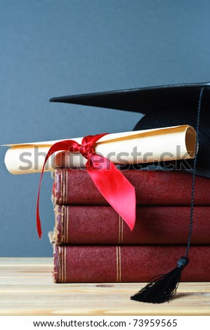 A stack of old, worn books with a mortarboard and ribbon tied scroll on top, placed on a wooden table with a grey background.