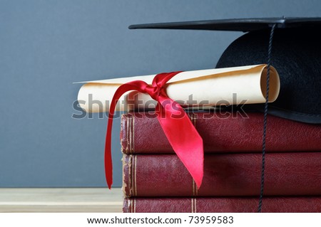 Close up of a mortarboard and graduation scroll on top of a pile of old, worn books, placed on a light wood table with a grey background.
