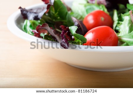A white bowl of salad lettuces and tomatoes, including red lettuce and rocket, on a light wooden table.  Landscape (horizontal) orientation.