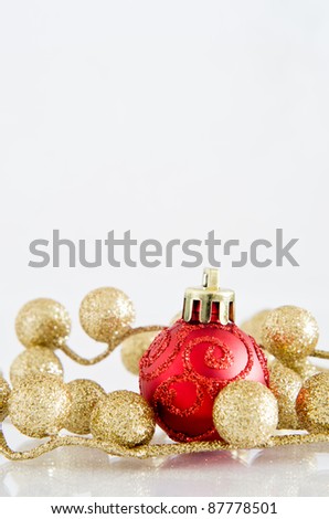 A red Christmas bauble with glittery spiral pattern and gold glitter ball decorations on an off-white reflective surface with copy space above.