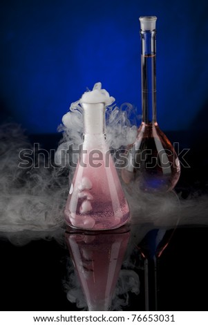 Vertical image of a pair of flasks, one with a red liquid, another with a bubbling pink liquid on a black reflective surface with a blue background.