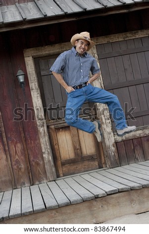 Angled vertical image of an Asian man in country garb kicking his heels in the air, as he jumps / dances in front of a western backdrop.