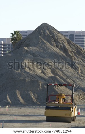 Vertical image of a steam roller dwarfed by a giant mount of asphalt at a construction site.