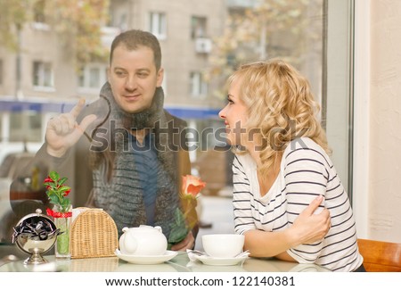 Date. Attractive young blond woman and her boyfriend at small cafe. Guy with rose draws a heart on the glass.