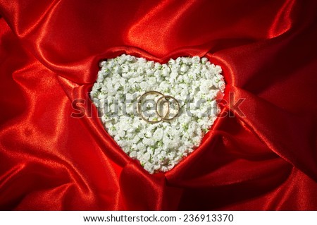 Marriage proposal background with red satin and heart shaped box under it. Two gold wedding rings on small white flowers.