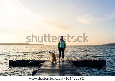 Woman with dog enjoy sunrise and lake, relaxing on bridge. Hiker or tourist looking at beautiful morning view with dog friend, inspirational landscape on beach. Peaceful people and serene concept.